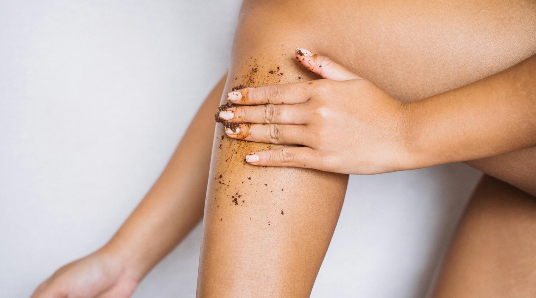 The image focuses on a close-up view of a person’s legs and hands, showcasing a body scrub application. The skin tone is a warm, medium brown, appearing smooth and healthy. The person’s hands are gently cradling one of their thighs, with visible traces of a dark brown, grainy scrub substance coating both the hands and the leg. This scrub appears to be a homemade or natural product, possibly containing coffee grounds or sugar, judging by the texture. The scrub is not uniformly applied; it’s concentrated in patches, suggesting the process of massaging it into the skin. The background is a clean, bright white, which emphasizes the contrast with the skin tone and the dark scrub. The lighting is soft and diffused, highlighting the texture of the skin and the scrub without harsh shadows. The composition is intimate and focuses on self-care and body positivity. The person’s posture and the gentle touch of their hands convey a sense of relaxation and pampering. The image evokes a feeling of wellness and natural beauty, suggesting a home spa treatment or a focus on skincare rituals. The nails are neatly manicured with a neutral polish, adding to the overall aesthetic of care and attention to detail.
