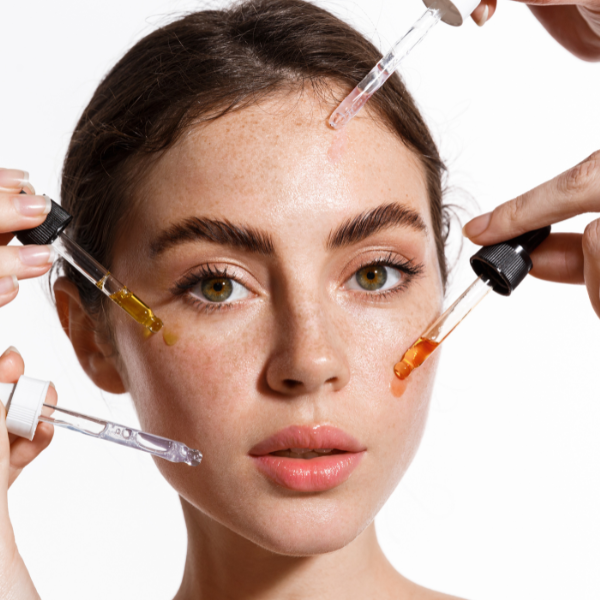 Close-up of a young woman with natural makeup applying multiple serums to her flawless skin, highlighting a beauty routine with clear, detailed droppers against a bright background.
