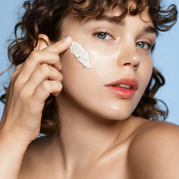 Radiant young woman with curly hair applying sunscreen on her cheek, showcasing a healthy skincare routine with a focus on sun protection against a soft blue background.