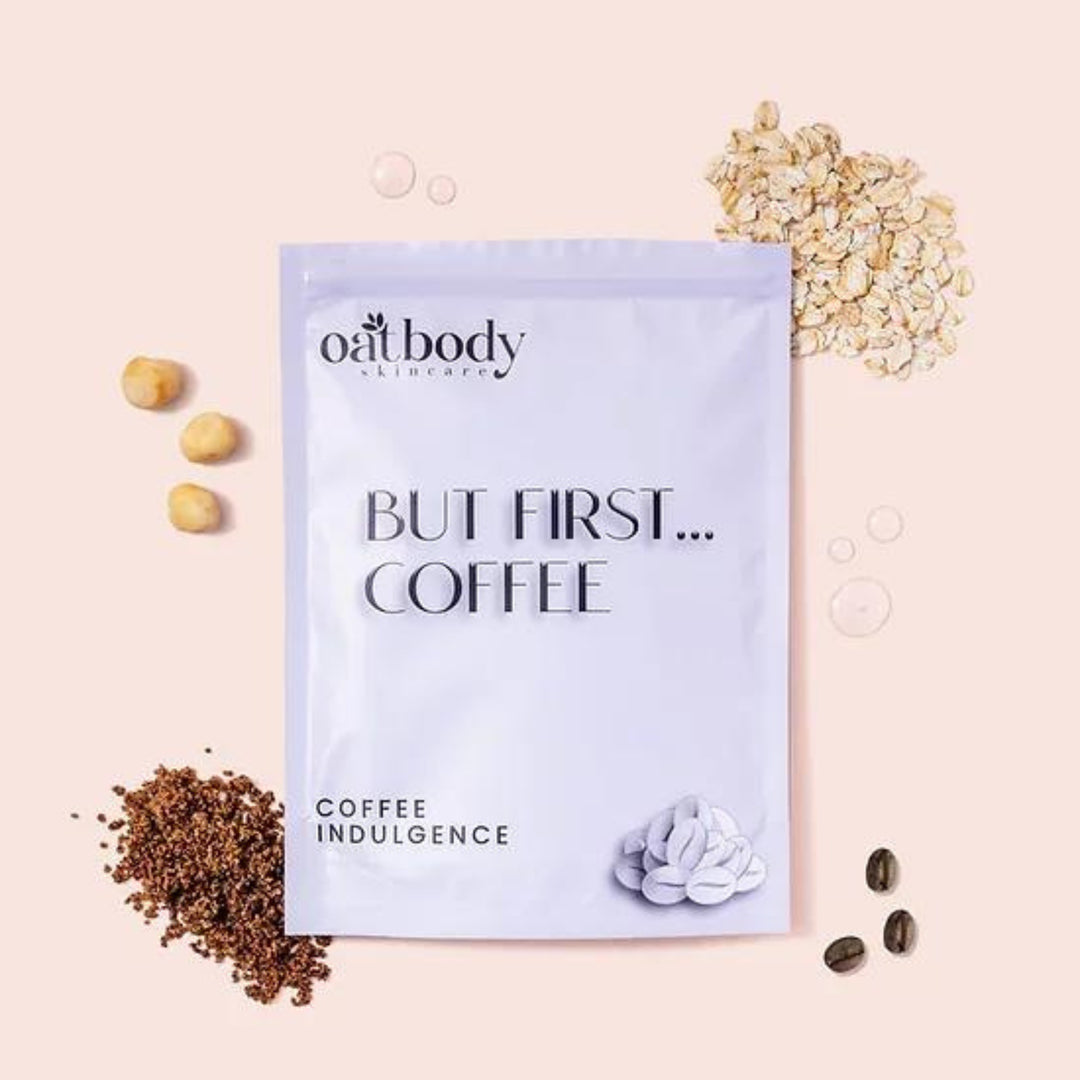 A bag of Oatbody Skincare's 'Coffee Indulgence' body scrub packaging with 'BUT FIRST… COFFEE' slogan on a peachy background, flanked by natural ingredients like ground coffee, oats, macadamia nuts. The product is a gentle body scrub, this product help with bumpy, rough, and dry skin. 