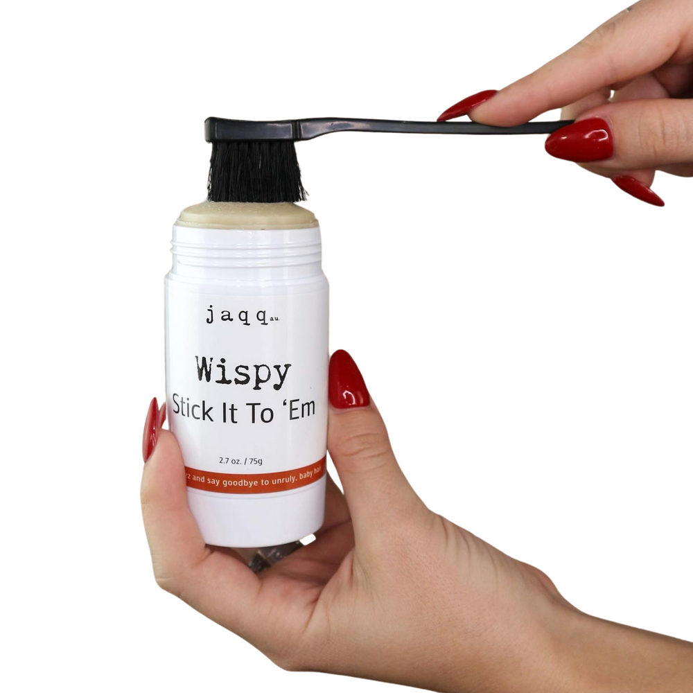 The image shows a person's hands holding the "Wispy Stick It To 'Em" hair wax product. The container's label clearly displays the product name "Wispy Stick It To 'Em" in bold, along with the weight "2.7 oz / 75g" beneath. The slogan "fight frizz and say goodbye to unruly, baby hair" is visible at the bottom. The other hand holds a small black brush with bristles, positioned over the open container, ready to apply the product, which is presumably a solid or semi-solid stick form. 
