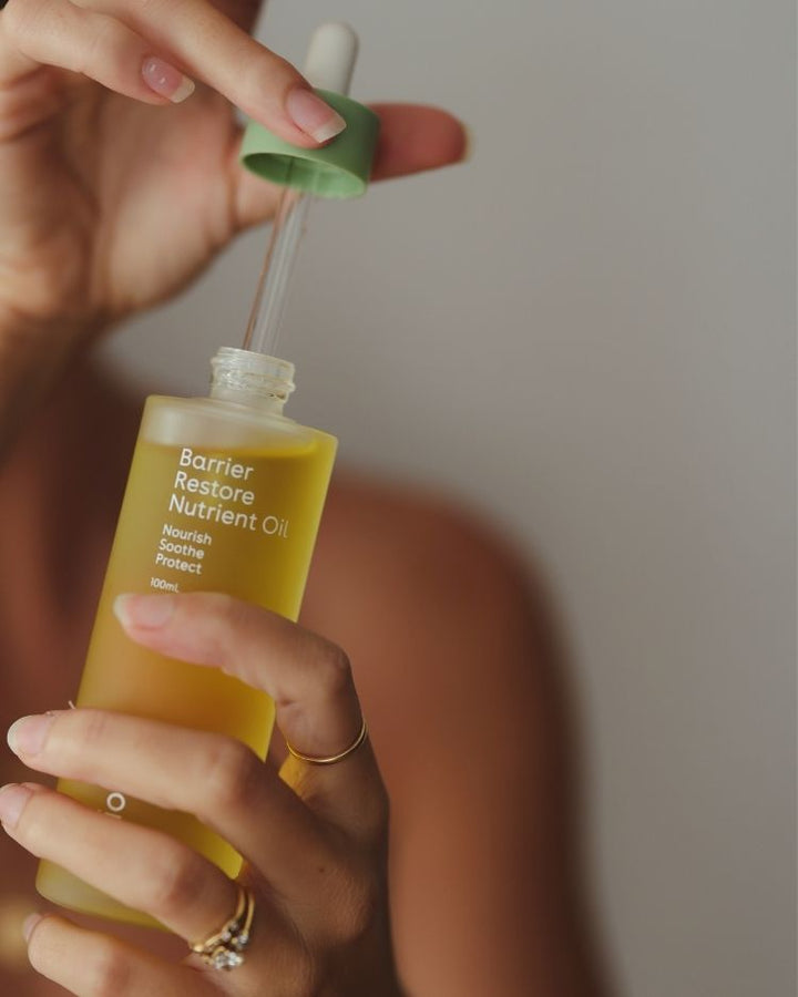 Hand holding a bottle of Barrier Restore Nutrient Oil with a dropper against a neutral background, Face oil for acne prone skin