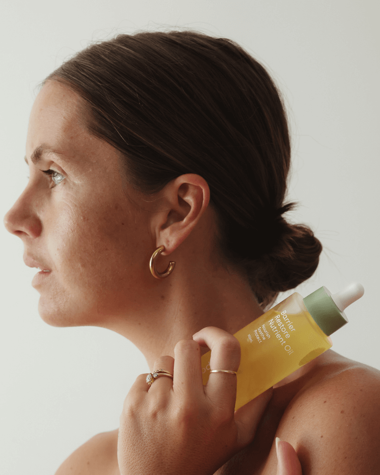 Woman applying a yellow face oil for acne prone skin to her neck with a neutral background.
