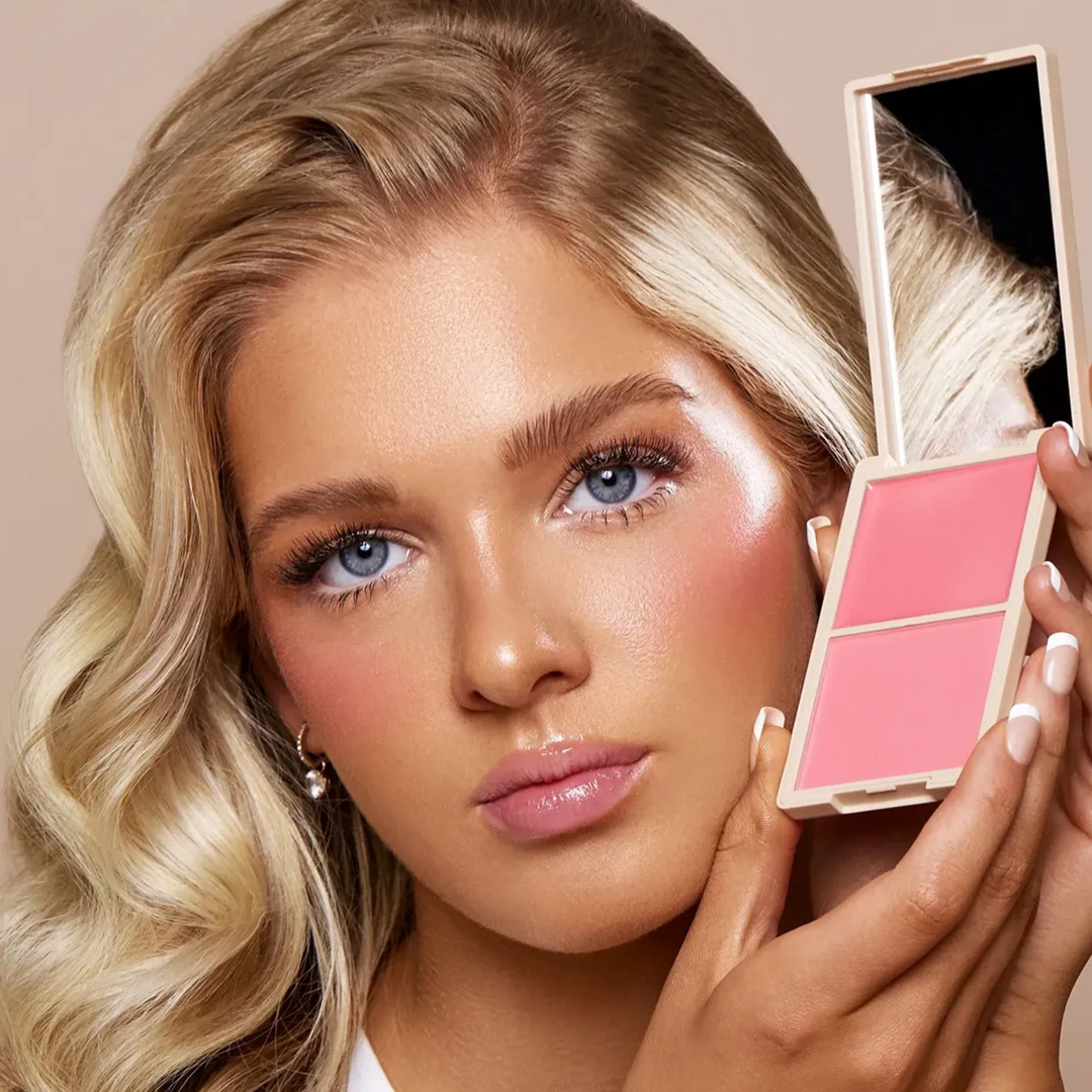 Model showcasing the Make Me Blush Cream & Powder Duo in the shade 'Sweet Cheeks,' holding the compact to highlight its vibrant pink tones. The duo creates a radiant, natural flush, complementing the model's glowing complexion and soft, glamorous makeup look.