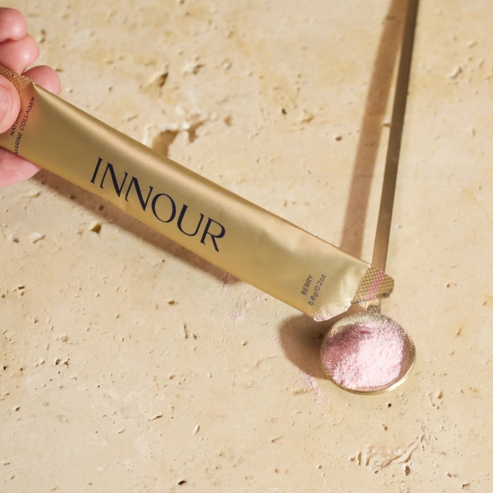 INNOUR Natural Marine Collagen sachet in Berry flavour being poured into a golden spoon. The fine pink collagen powder, with 5000mg per serve, is formulated to enhance skin hydration, elasticity, and overall wellness, showcased against a textured stone surface.