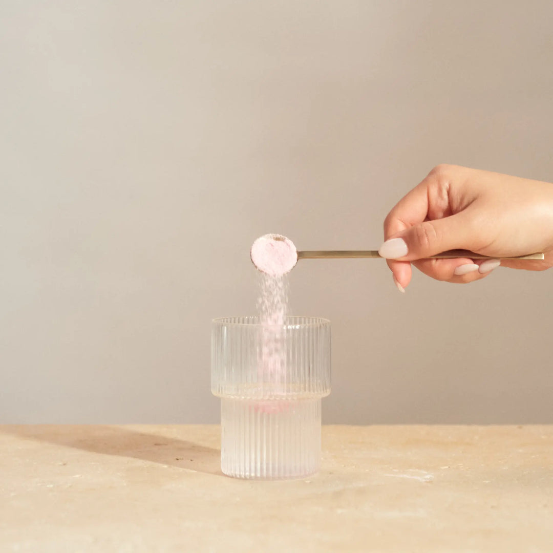 Hand using a small scoop to add marine collagen powder into a clear glass container on a wooden surface.