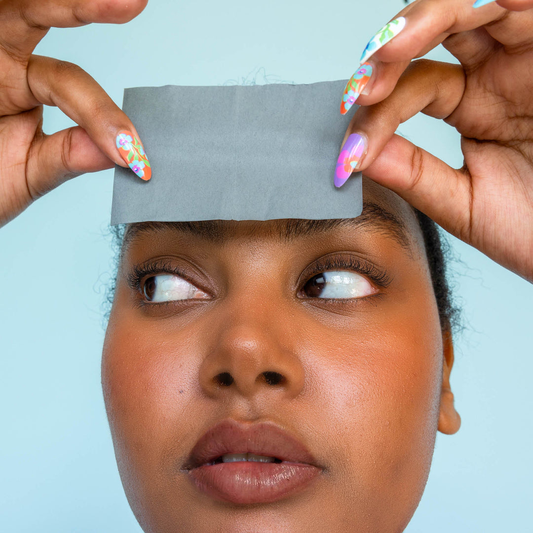 Model using Skoop Skincare Babe Blotter Extra Drip Charcoal Blotting Paper on her forehead, demonstrating its oil-absorbing effectiveness. The close-up highlights her glowing skin and colourful nail art, set against a soft blue background, showcasing a fresh and vibrant skincare routine.