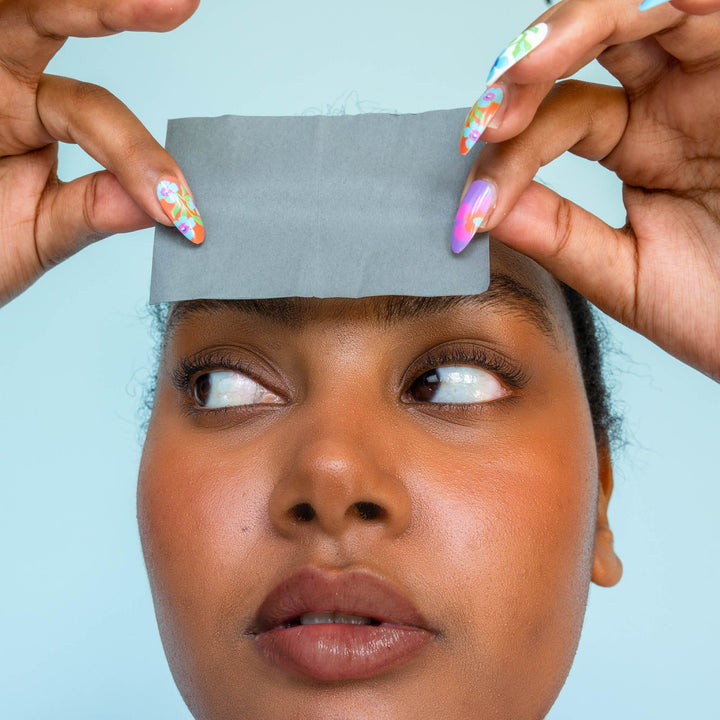 Model using Skoop Skincare Babe Blotter Extra Drip Charcoal Blotting Paper on her forehead, demonstrating its oil-absorbing effectiveness. The close-up highlights her glowing skin and colourful nail art, set against a soft blue background, showcasing a fresh and vibrant skincare routine.