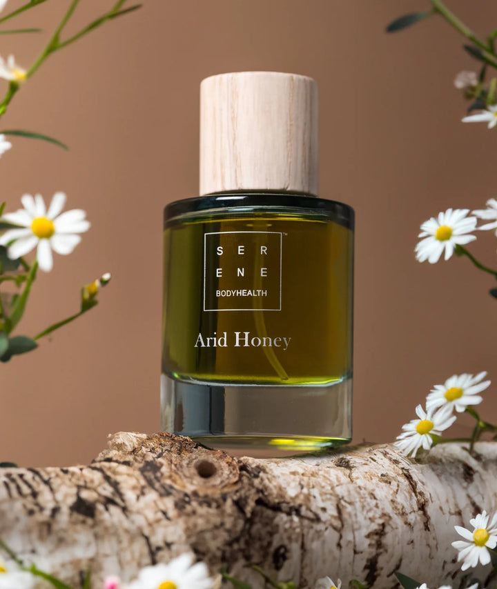 Arid Honey fragrance bottle by Serene Bodyhealth displayed on a birch log surrounded by delicate white flowers.