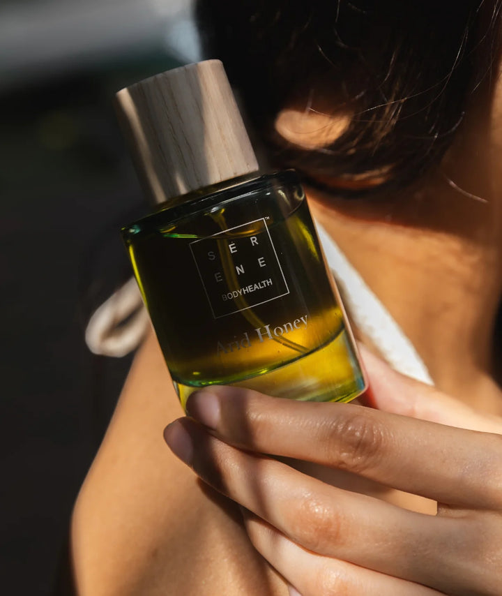 Close-up of a woman holding Serene Bodyhealth's Arid Honey perfume bottle in sunlight.