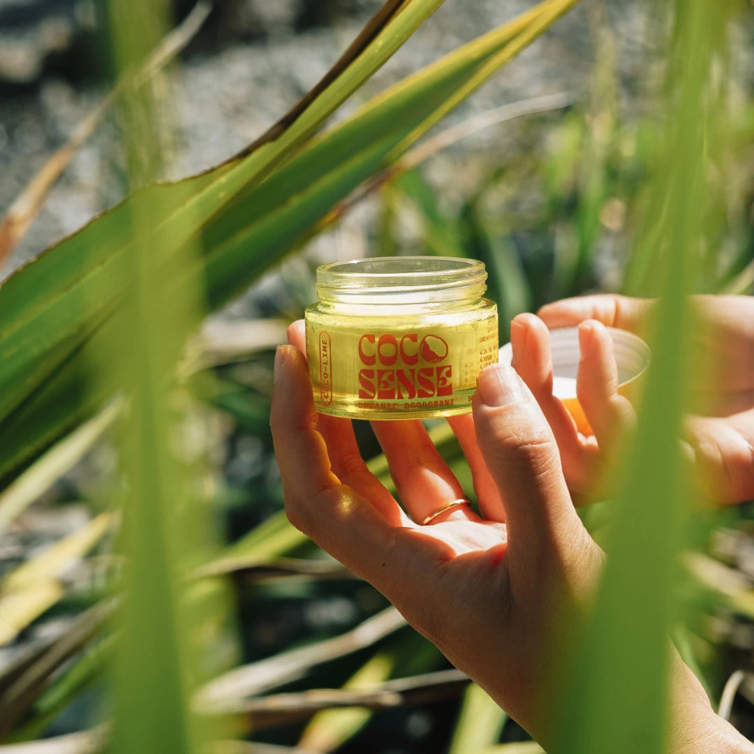 Hand holding a jar of best organic deodorant labeled 'Good Sense' amidst green leaves