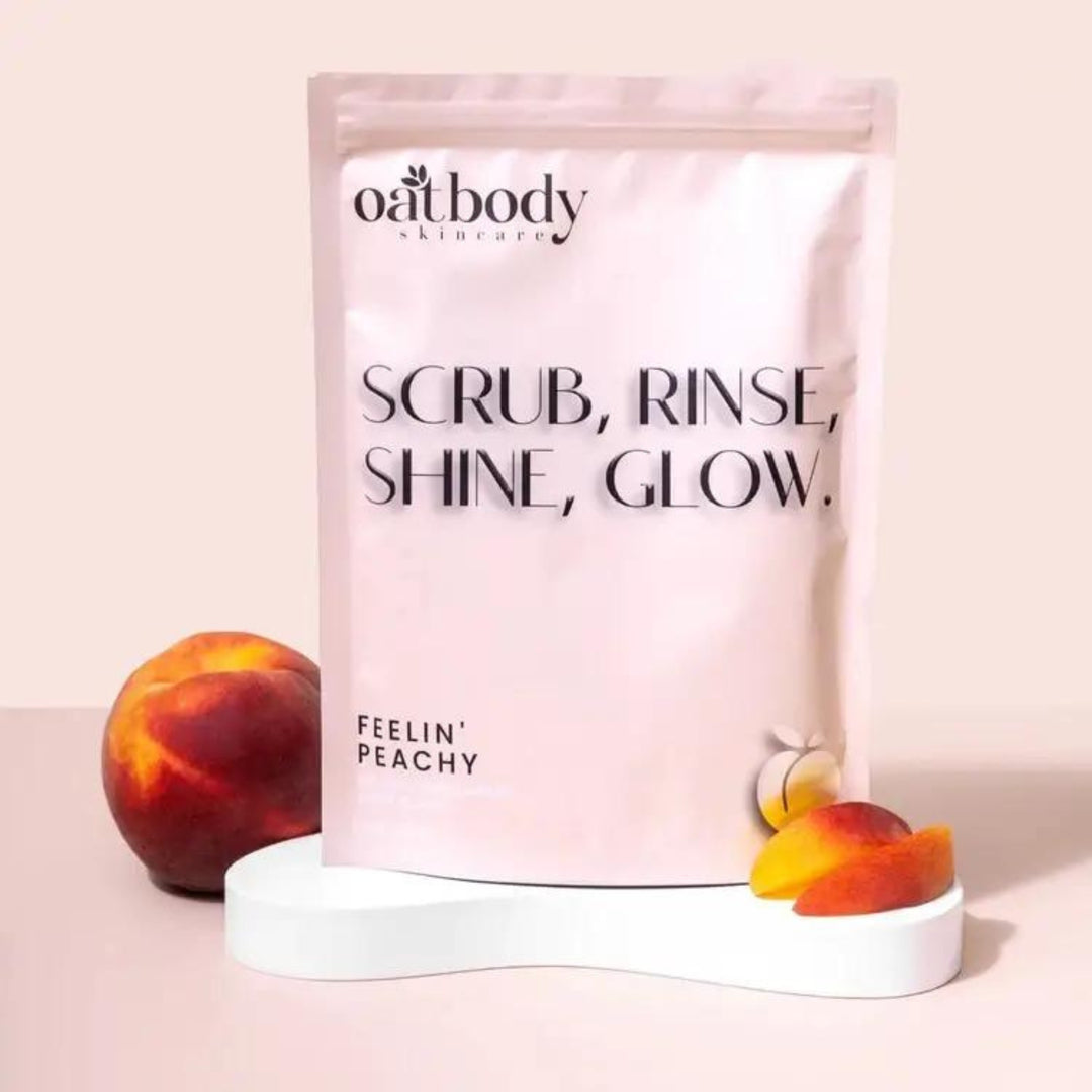 Oatbody Skincare's 'Feelin' Peachy' body scrub package with a catchy phrase 'SCRUB, RINSE, SHINE, GLOW.' displayed alongside a fresh peach on a modern, neutral-toned background. The product is a gentle body scrub, this product helps with bumpy, rough, dry skin, strawberry skin, and keratosis pillaris.