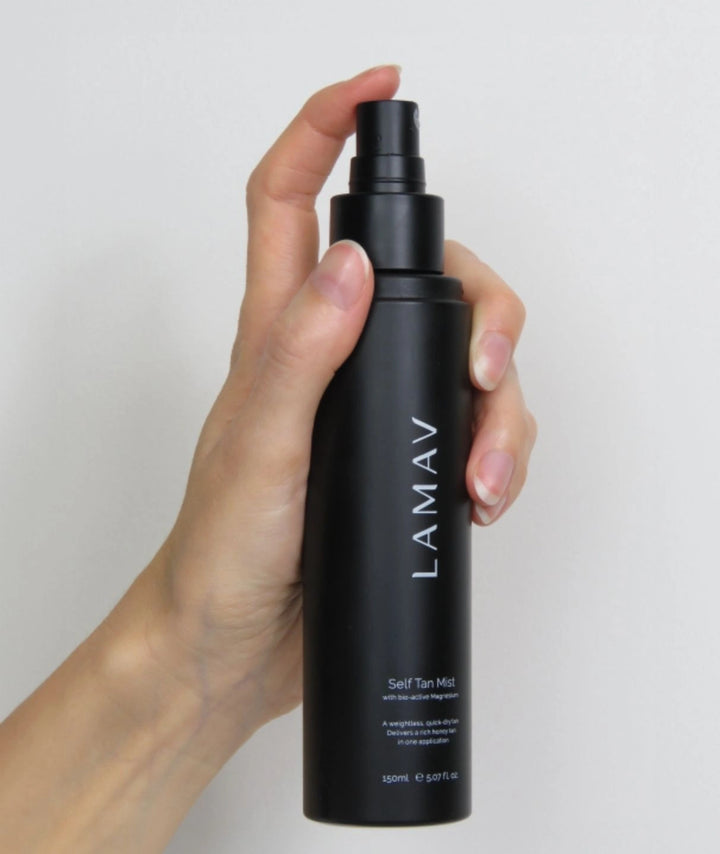Hand holding and spraying the LAMAV Self Tan Mist bottle against a white background.