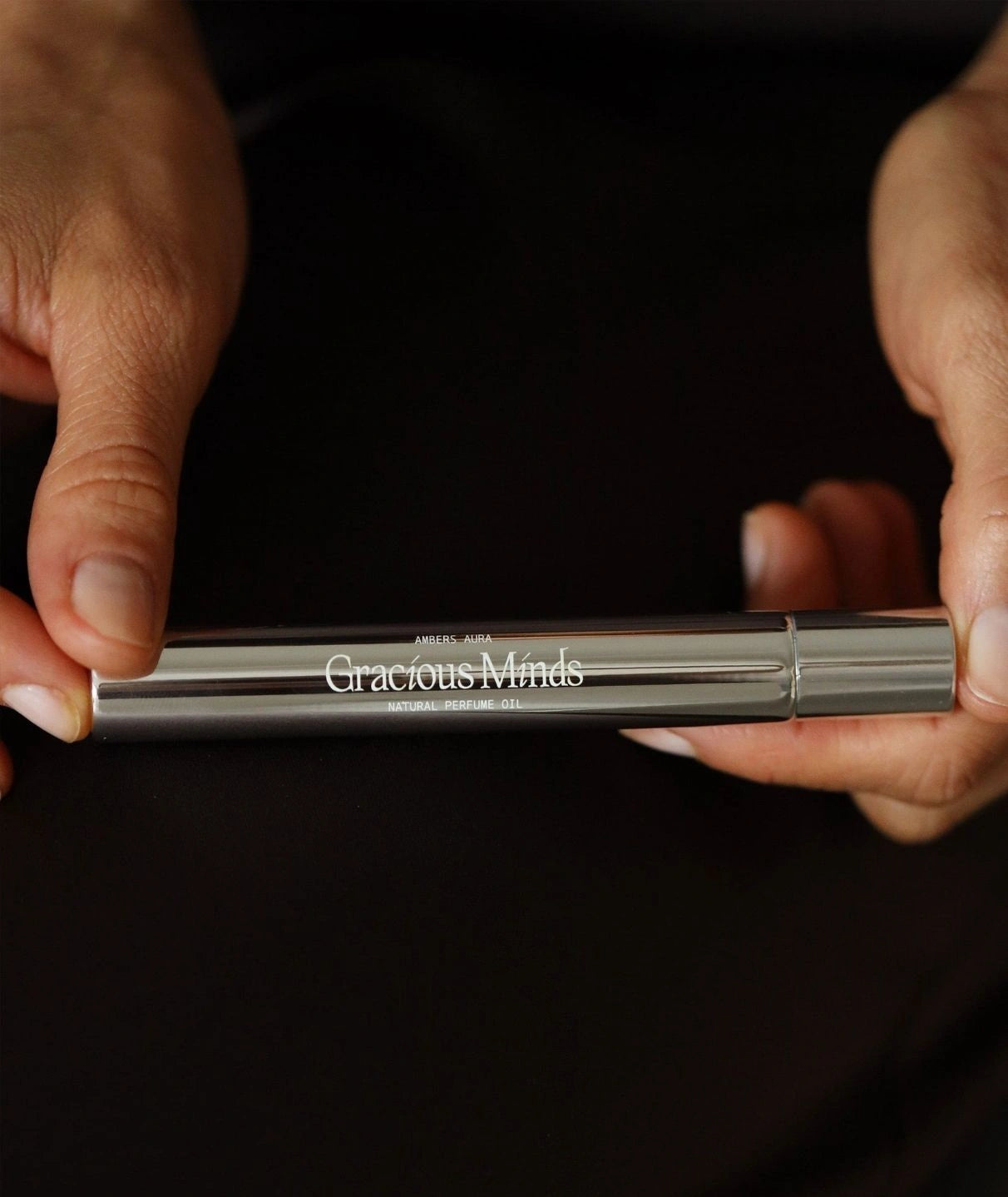 Hands holding the metallic Gracious Minds Ambers Aura natural perfume oil roller.