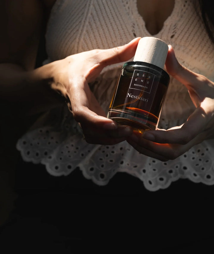 Woman holding Serene Bodyhealth Nestinari Eau de Parfum bottle with a wooden cap in soft sunlight.