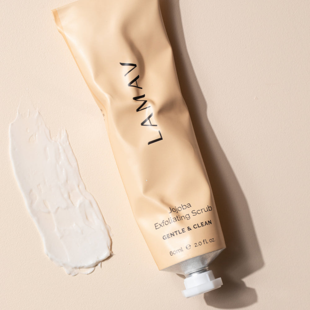 A tube of LAMAV Jojoba Exfoliating Scrub is lying diagonally next to a smeared sample of the product on a light-colored surface. The beige tube with black text specifies that it's a 'GENTLE & CLEAN' exfoliant, 60ml or 2.0 fl oz. The creamy white texture of the scrub is visibly thick and spread out, indicating the product's consistency and use.