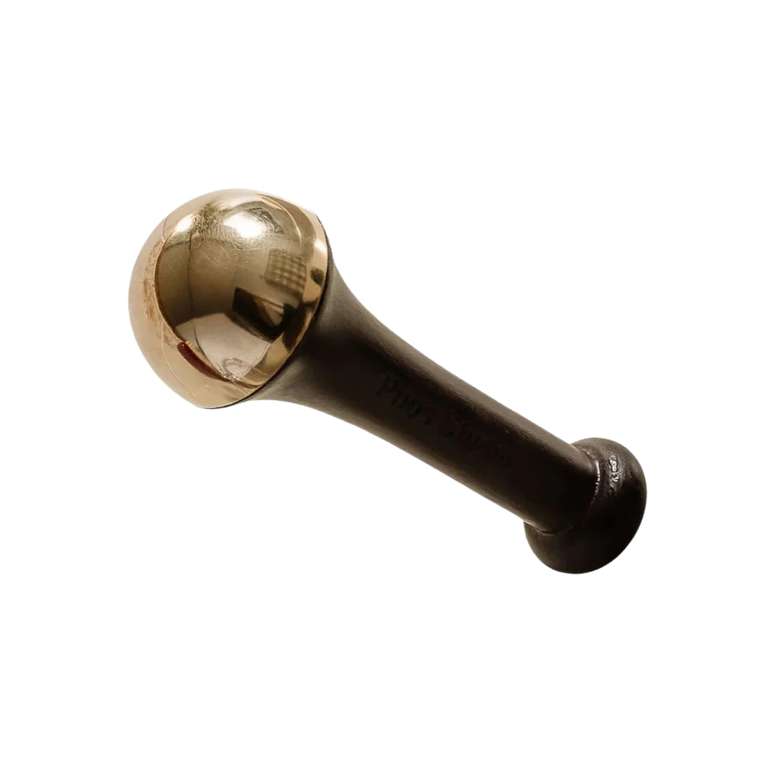 A close-up of a Kansa wand, an Ayurvedic massage tool, with a polished bronze dome and dark wooden handle. The wand is set against a plain background, highlighting its reflective metal head used for rejuvenating facial massages.