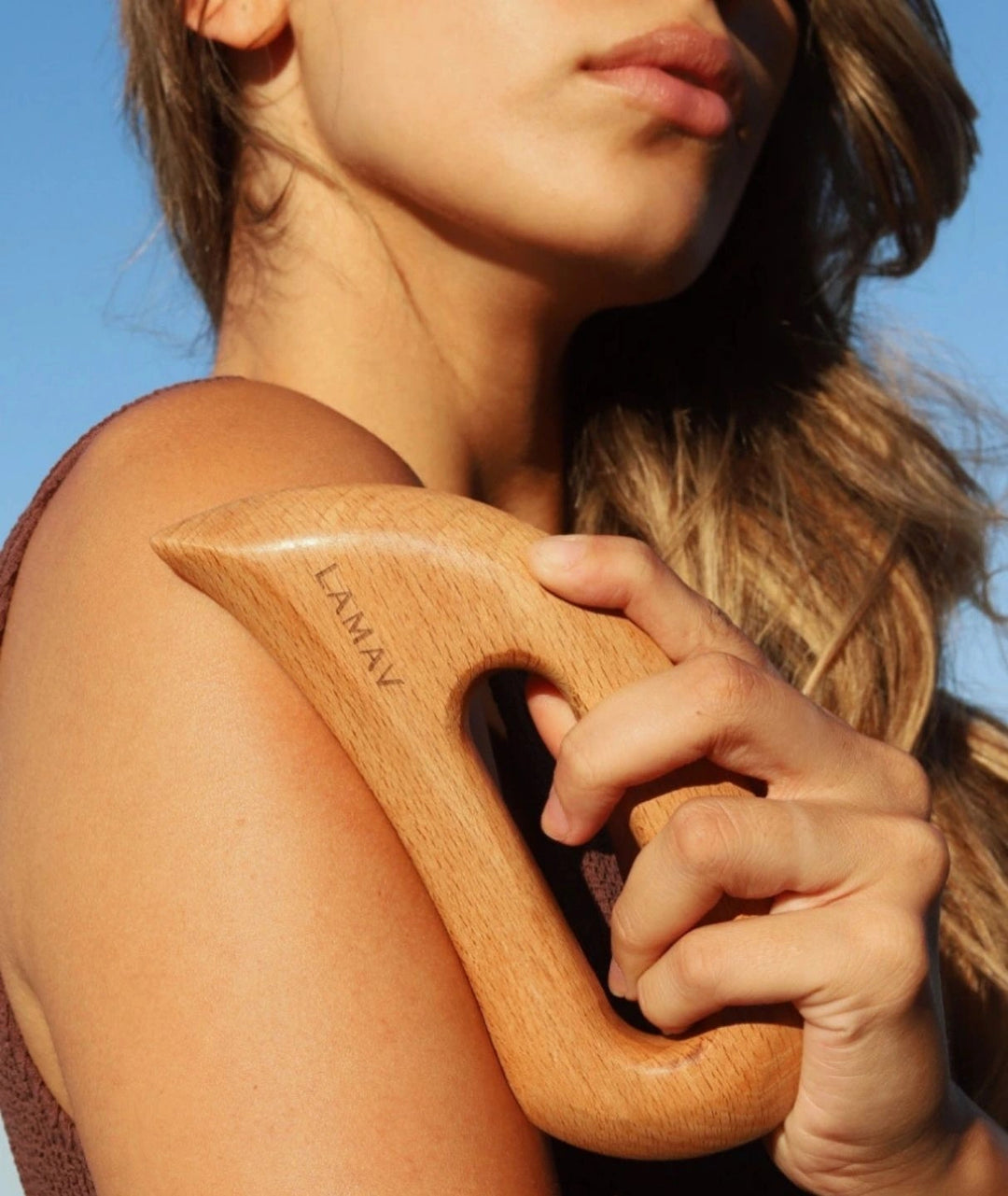 Woman outdoors using the LAMAV Aura Body Gua Sha on her shoulder for lymphatic massage.