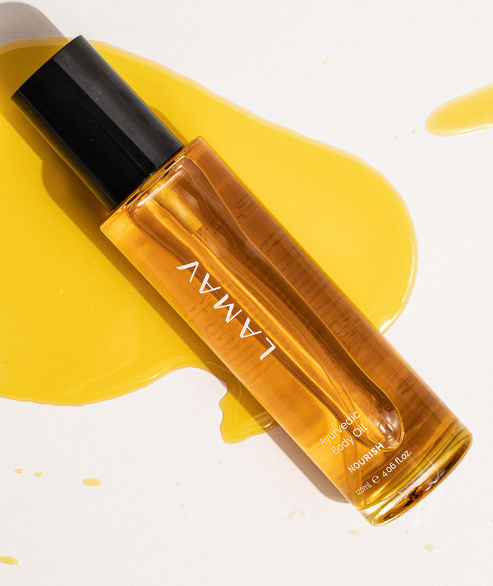 LAMAV Ayurvedic Body Oil Nourish bottle lying on a smear of golden oil, showing product texture.