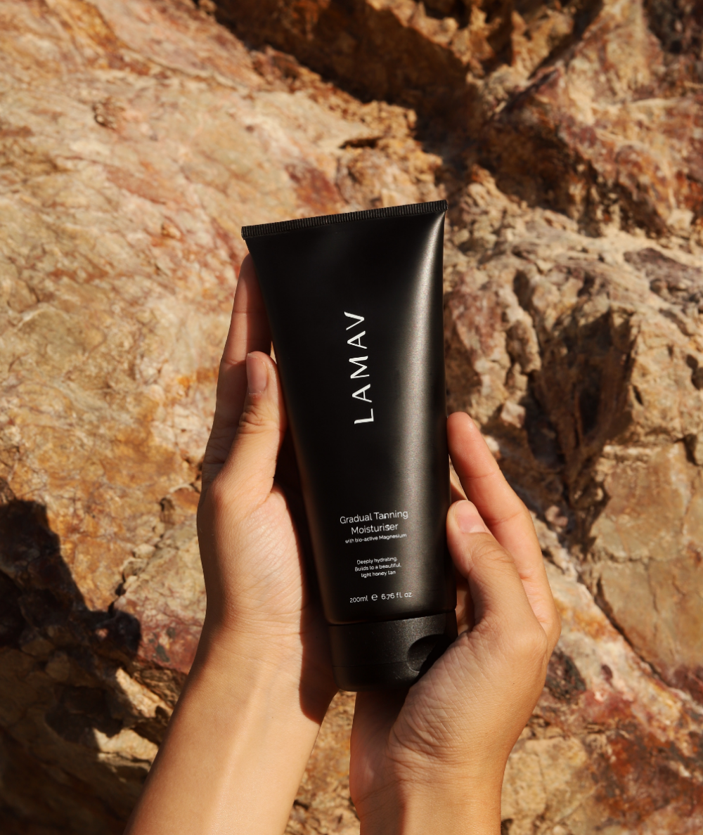 Hands holding LAMAV Gradual Tanning Moisturiser outdoors against natural rocks. Hydrating sunless tanner.