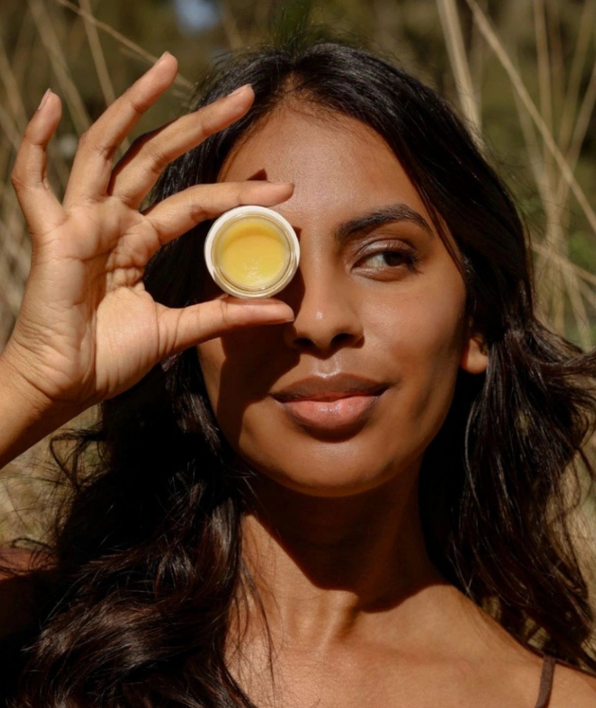 Woman outdoors holding an open jar of LAMAV Intense Repair Eye Balm over her eye in natural sunlight.