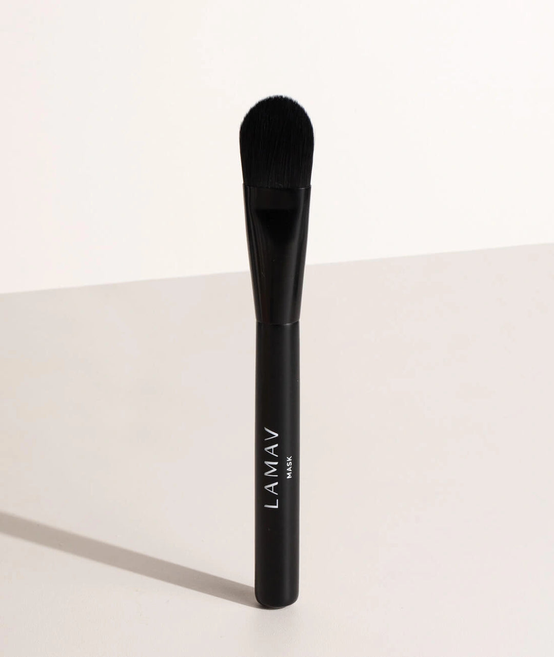 Standing LAMAV Vegan Mask applicator brush on a soft beige surface.