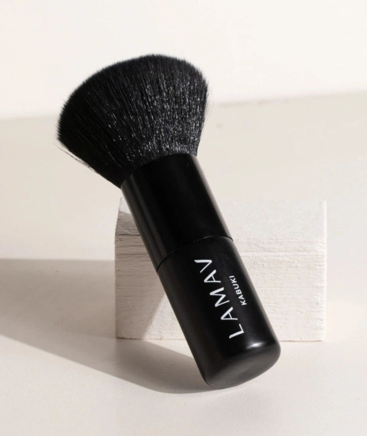 LAMAV Vegan Kabuki Brush leaning against a white textured block. Premium black makeup applicator tool.