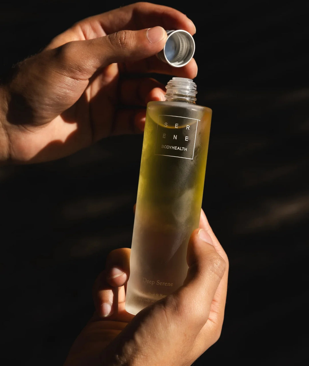 Hands opening a bottle of Serene Bodyhealth Deep Serene magnesium body oil in natural light.