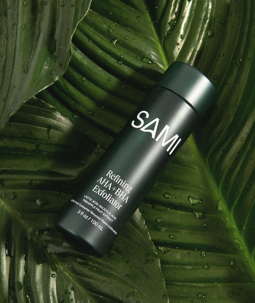 Refining AHA BHA Exfoliator by SAMI — black bottle lying on large wet green leaves | Natural botanical exfoliation