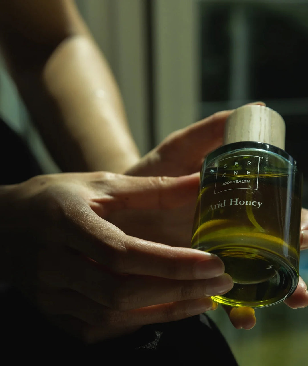 Hands holding the Arid Honey glass perfume bottle by Serene Bodyhealth in soft ambient lighting.