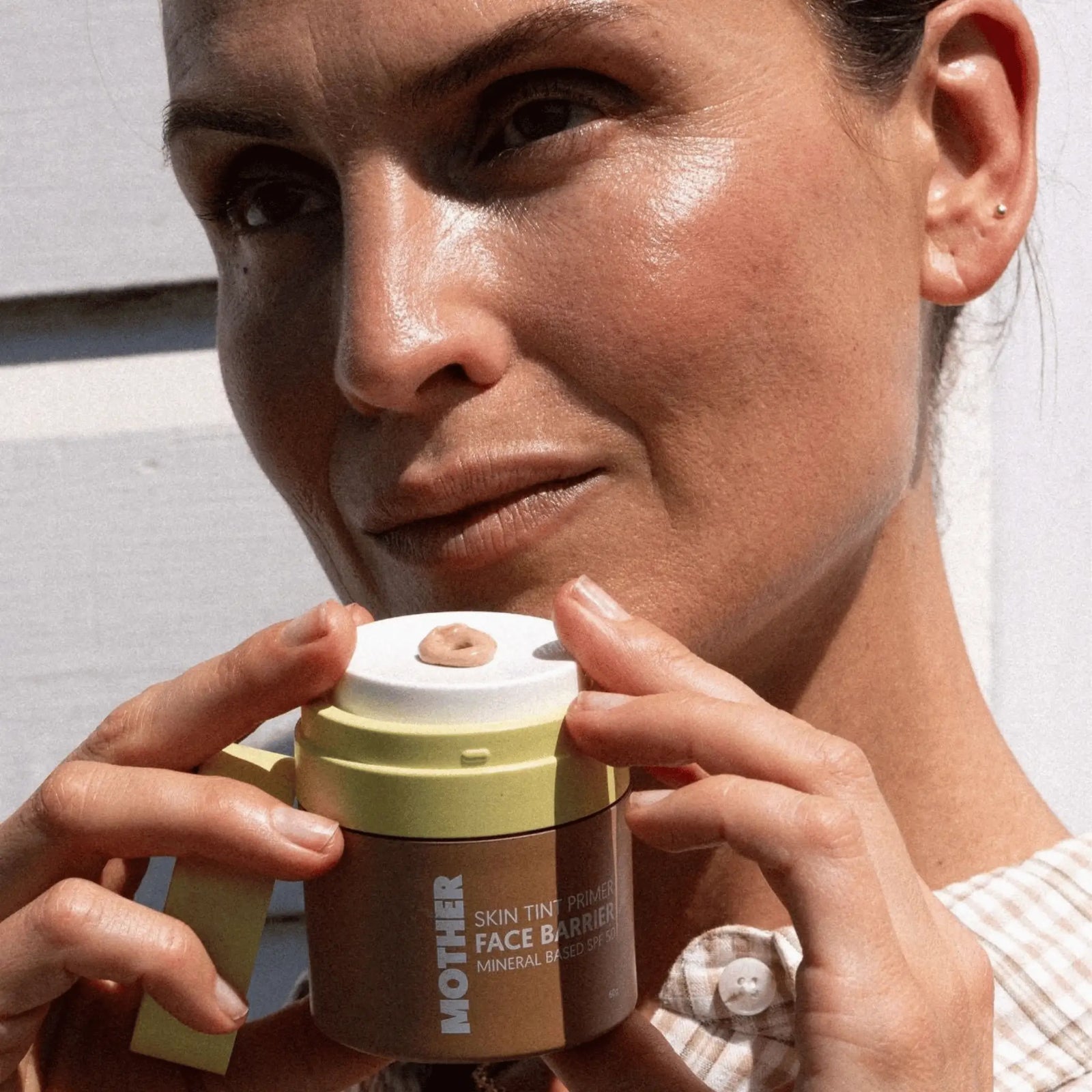 Close-up of woman holding Mother SPF Skin Tint Face Barrier, a mineral sunscreen and primer for oily acne prone skin.
