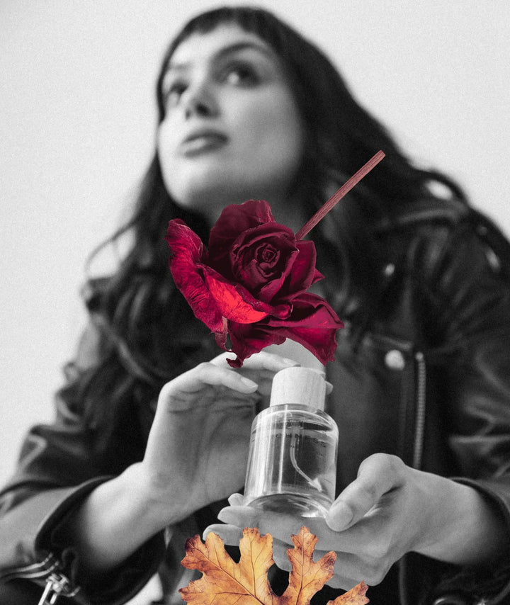 Woman holding Suchness perfume bottle by Serene Bodyhealth, styled with a deep red rose and autumn leaf.