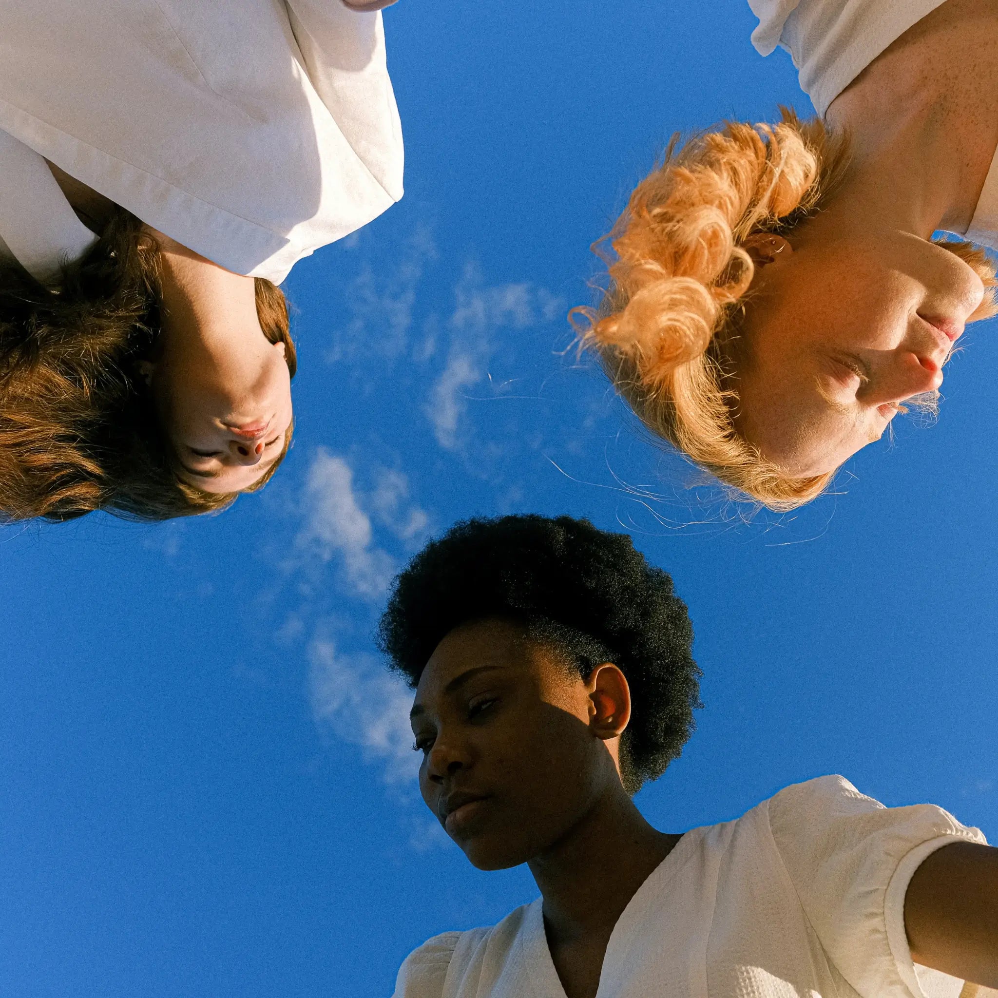 A low-angle shot of three diverse women standing against a clear blue sky, looking down into the camera, creating an empowering perspective. They are holding hands in a show of unity. The woman in the foreground has a short afro, the second has long, wavy dark hair, and the third has curly blonde hair. They all wear white, suggesting a theme of purity, peace, or solidarity.