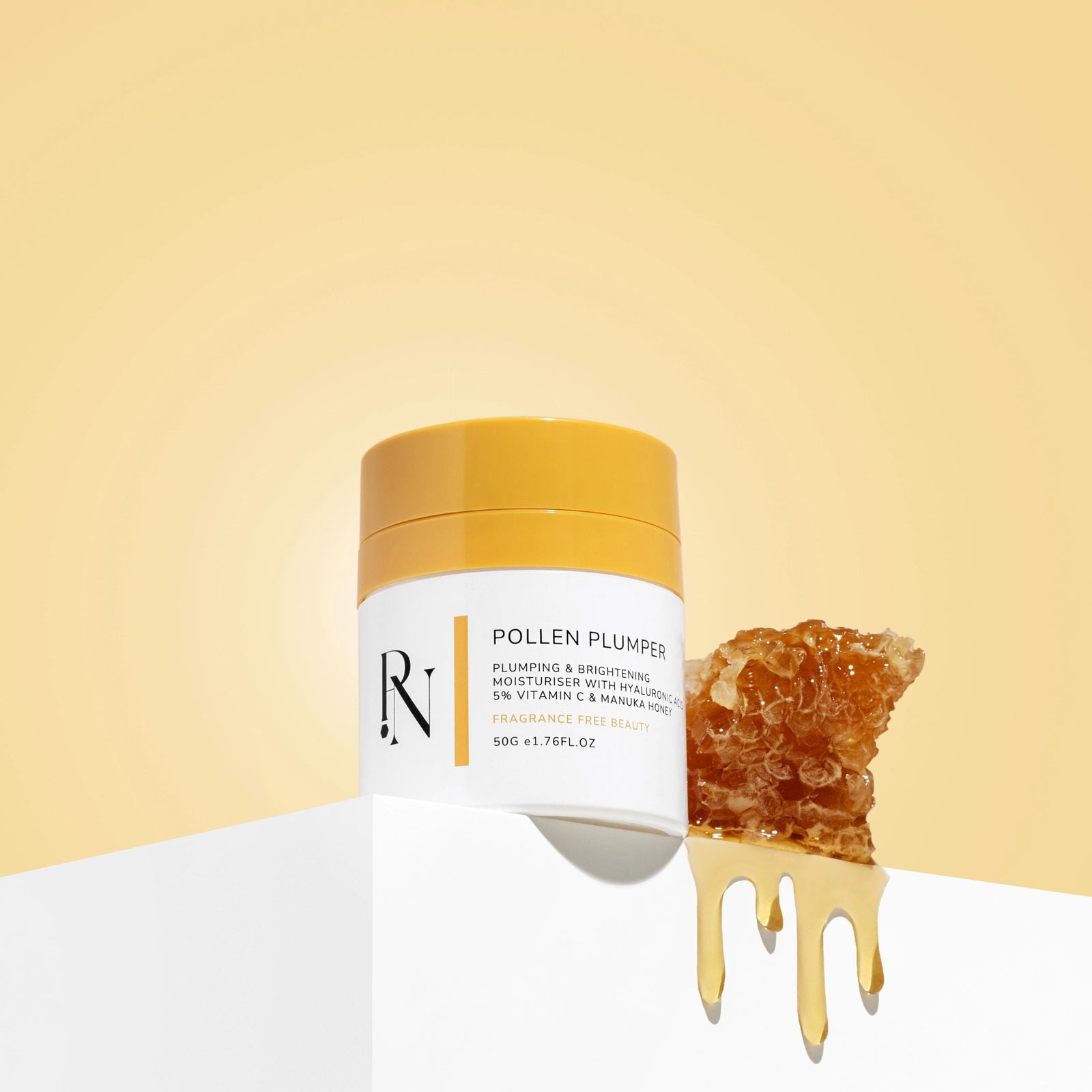 A jar of Pollen Plumper moisturizing and brightening facial moisturiser  from PollenNation Skin rests on a reflective white surface against a warm yellow background. To the right, a piece of honeycomb drips with golden honey, suggesting the natural ingredients used in the cream. The product is 50g or 1.76 fl oz and contains hyaluronic acid and 5% Vitamin C with manuka honey.