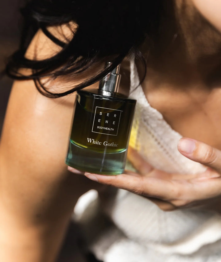 Woman holding the White Gothic fragrance bottle by Serene Bodyhealth against her shoulder in soft lighting.